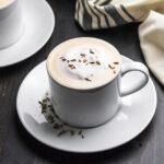 White mug of tea topped with frothed milk and lavender buds, sitting on white saucer.