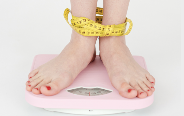 Bare feet standing on pink bathroom scale with yellow measuring tape wrapped around ankles.