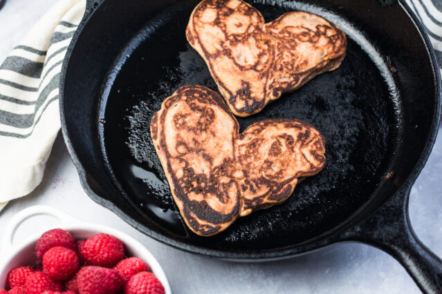 Two heart-shaped pancakes in a cast-iron skillet alongside a bowl of fresh raspberries.