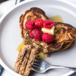 Heart-shaped pancakes topped with raspberries, butter, syrup on white plate with fork and blue striped napkin