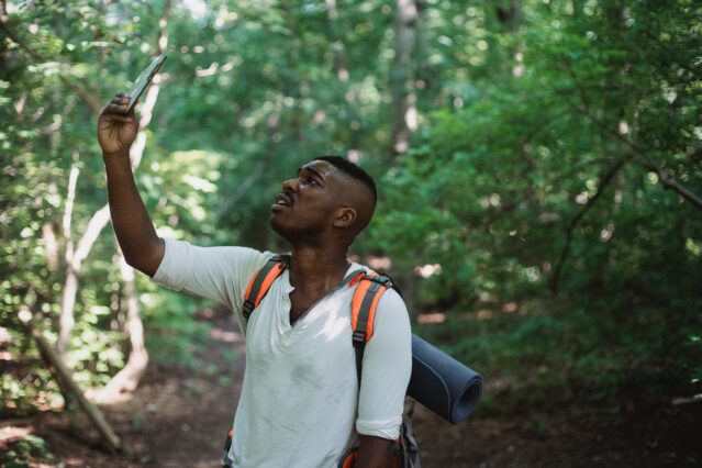 Man wearing hiking backpack in woods, holding up cell phone looking for reception