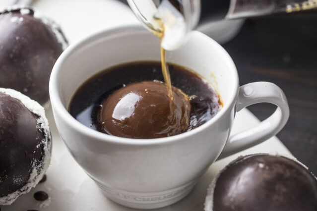 Chocolate bomb inside white mug of coffee with coffee pouring over top.