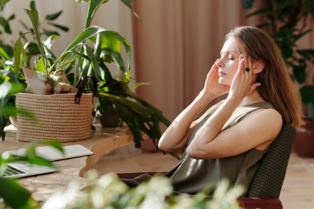 Woman sits in room surrounded by houseplants, eyes closed, rubbing temples
