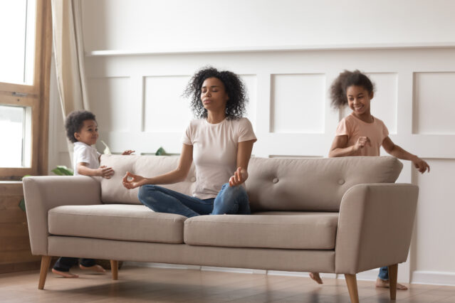 Mom sitting cross-legged on sofa in a meditation pose with two children running around the sofa