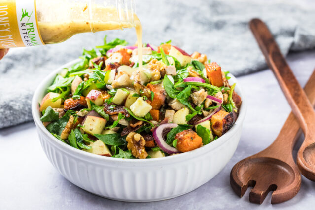 Warm spinach salad with cubed apples, butternut squash, walnuts, and sliced red onion, Primal Kitchen Honey Mustard being poured over top.