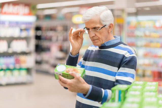 Older man in striped sweater stands in grocery store reading the label on a green box of food 