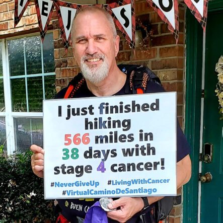 Smiling man holds a sign reading "I just finished hiking 566 miles in 38 days with stage 4 cancer."