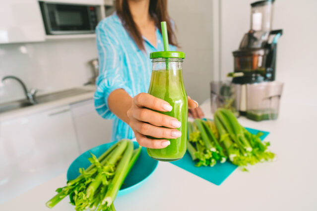 Woman in kitchen offering bottle of freshly pressed juice in juicer machine. 