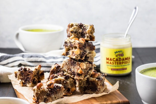 Blondie bars stacked on a cutting board. In background, cups of Primal Kitchen Matcha Lattes and a jar of Brad's Macadamia Masterpiece.