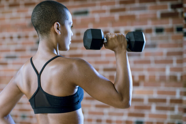 Woman wearing black sports bra is lifting dumbbell while facing a brick wall.