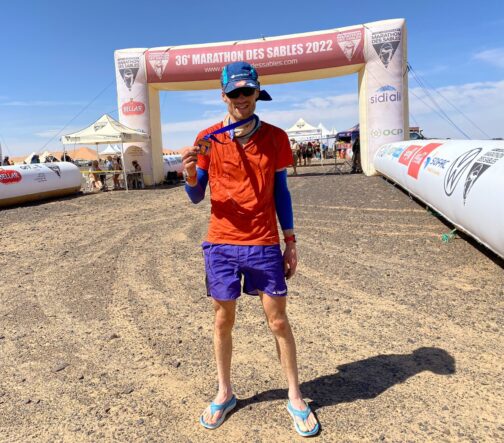 Man in red shirt and purple shorts stands in front of Marathon des Sables finish line showing off medal.