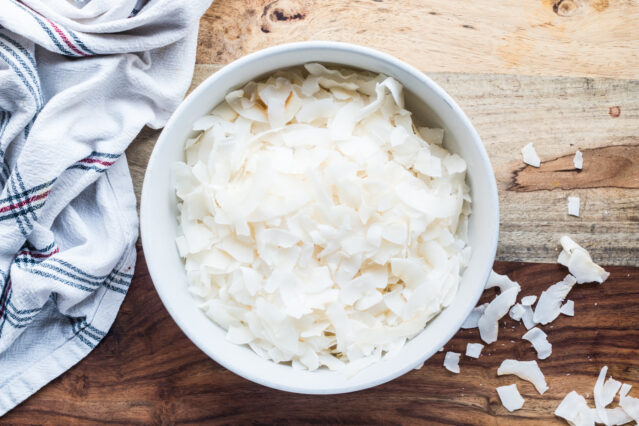 Bowl of large coconut flakes sitting on a wooden surface with a white kitchen towel.