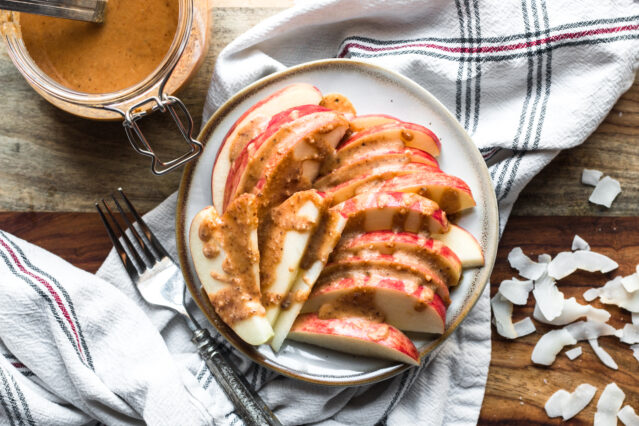 Apples on a plate drizzled with coconut butter alongside white kitchen towel, fork, jar of coconut butter, raw coconut flakes.