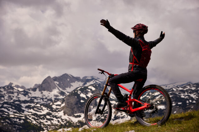 Male mountain biker stands with arms outstretched on mountaintop astride red bike