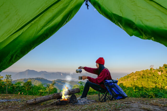 View from inside green tent of woman enjoying hot drink in front of campfire