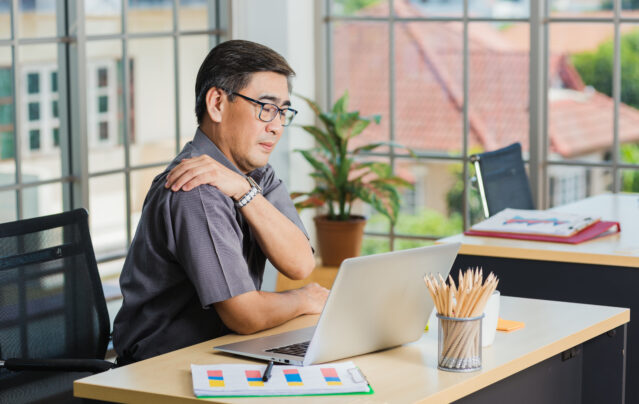 Man sits at desk with laptop rubbing shoulder.