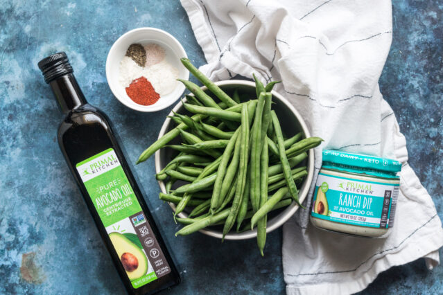 Bowl of fresh green beans, bowl of spices, Primal Kitchen Avocado Oil and Ranch Dip, white kitchen towel.