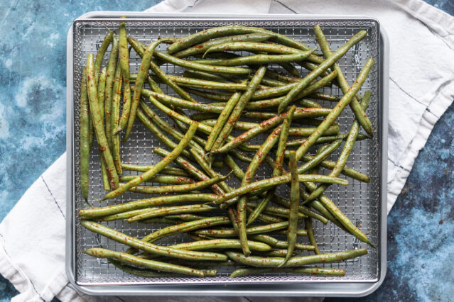 Spiced green beans in an air fryer basket atop white kitchen towel.