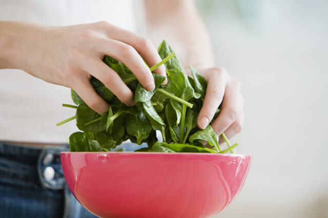 Hands tossing fresh spinach in a red bowl.