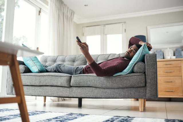 Shot of a man using a remote control while lying on the couch at home.