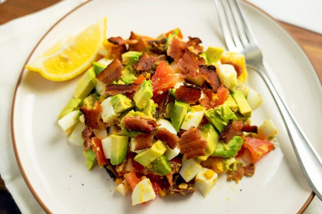 Bacon, egg, avocado, tomato salad on white plate with form and lemon wedge