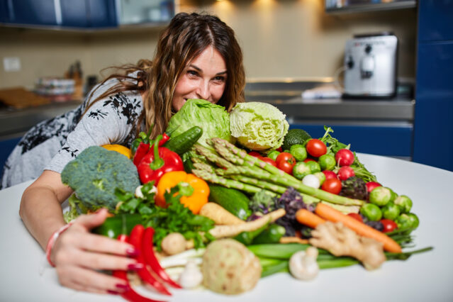 Woman embraces a big pile of assorted vegetables piled on a kitchen countertop.