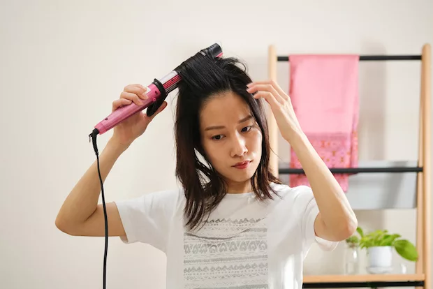 Asian woman curling her hair, which may cause dry hair and frizz
