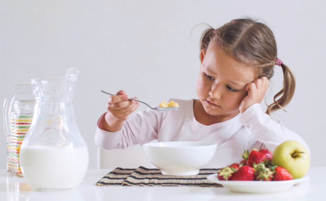 Girl looking sadly at bowl of cereal, with plate of fruit and pitcher of milk on the table.