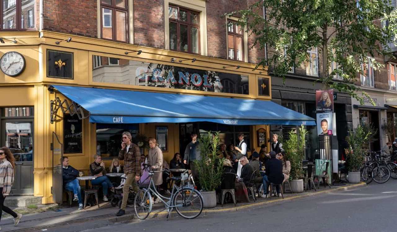 People walking by people seated at an outdoor cafe on a leafy street in the neighborhood of Vesterbro in Copenhagen, Denmark