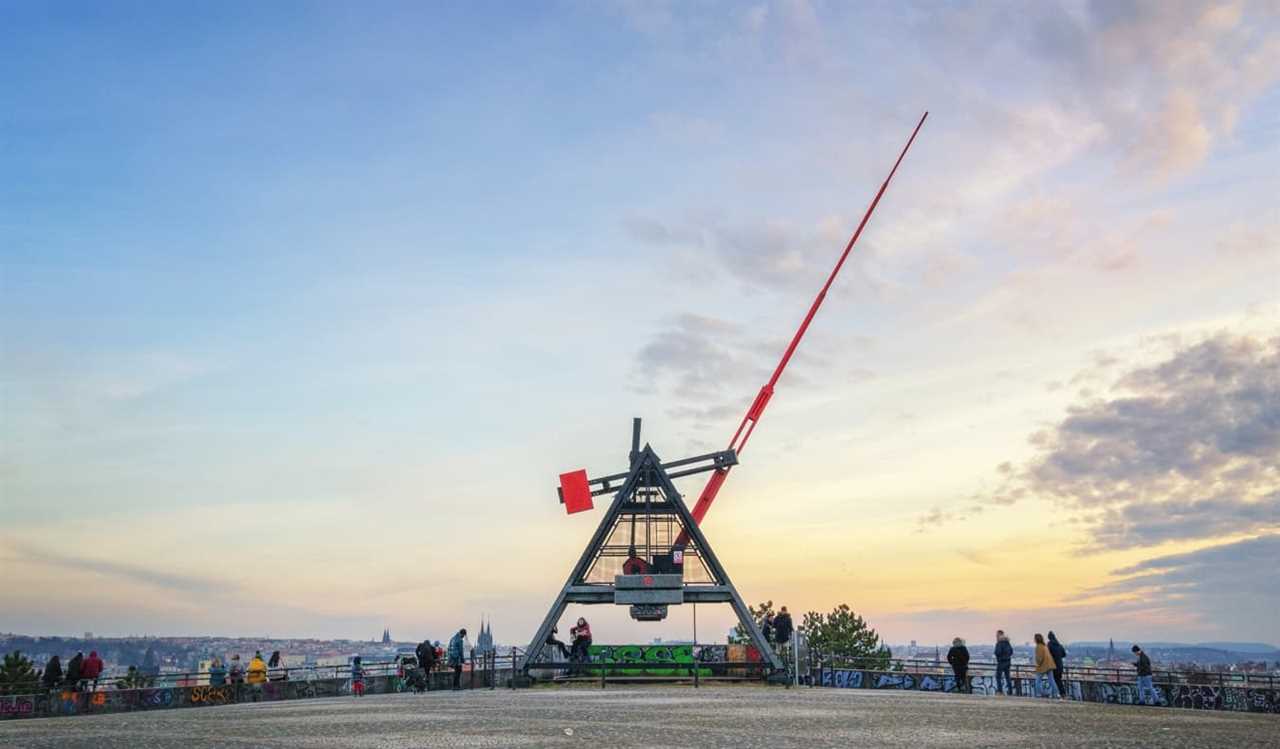 A giant metal metronome with the long red hand pointing into the sky at sunset, in Prague, Czech Republic