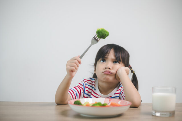Girl sits at a table with a bowl of vegetables looking skeptically at a piece of broccoli speared on her fork.