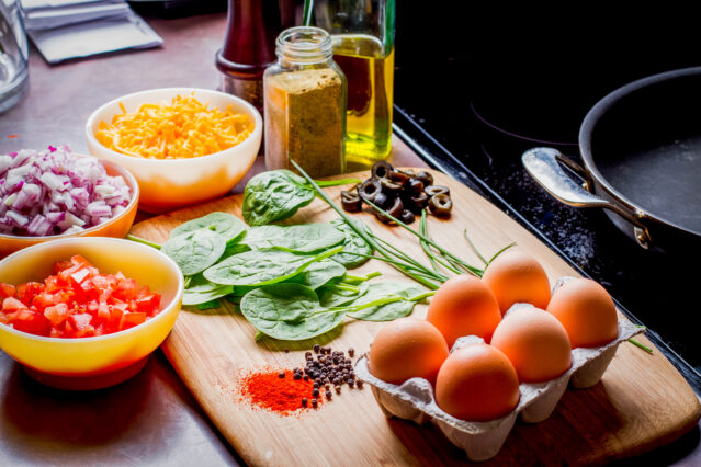 Eggs, cheese, spinach, onion, olives, tomato and seasonings on a bamboo cutting board.