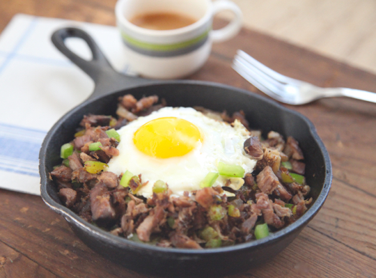 Shredded pork in cast iron skillet with fried egg on top, fork and mug of coffee in the background.