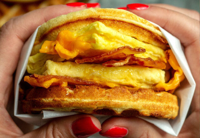 Woman's hands holding waffle sandwich with eggs and bacon.