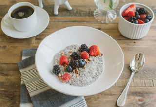 White bowl of chia pudding with berries on table with coffee, bowl of berries, napkin, and fork.