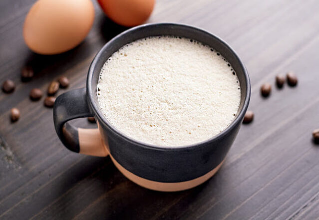 Mug of frothy egg coffee on a dark wood table with eggs and coffee beans.