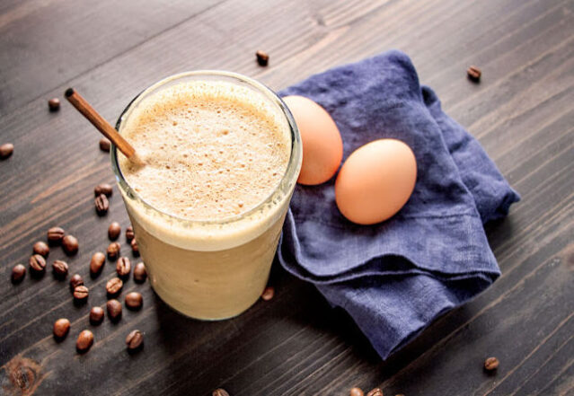 Glass of frappe with a straw on a dark wood table next to blue napkin with two eggs on top.