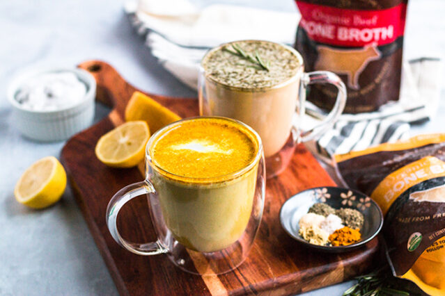 Two glass mugs of bone broth lattes on top of a dish towel surrounded by various spices and lemon slices.