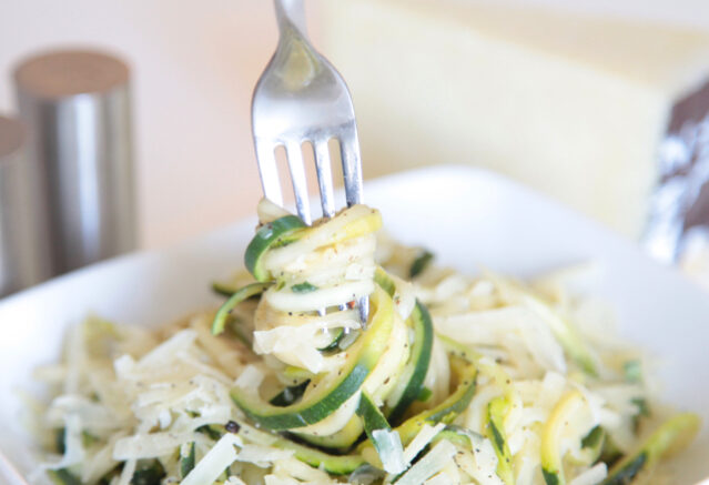 A fork swirling zucchini noodles with shredded cheese out of a white bowl.