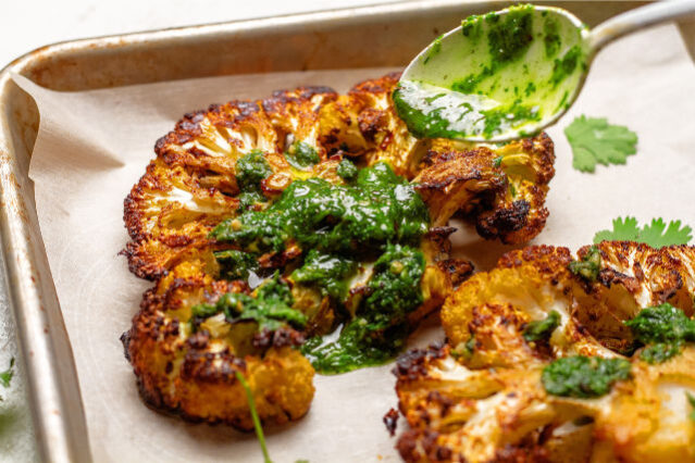 Spoon drizzling bright green chimichurri sauce over a roasted cauliflower steak on a sheet pan lined with parchment paper.