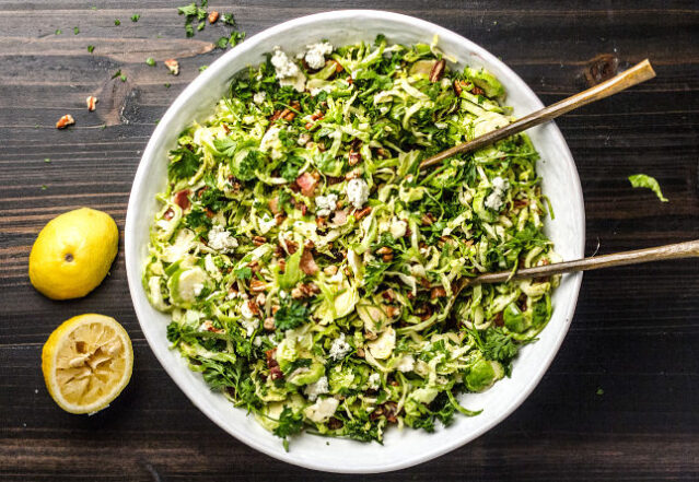 Overhead shot of shaved Brussels sprouts salad in large white serving bowl with two lemon halves.