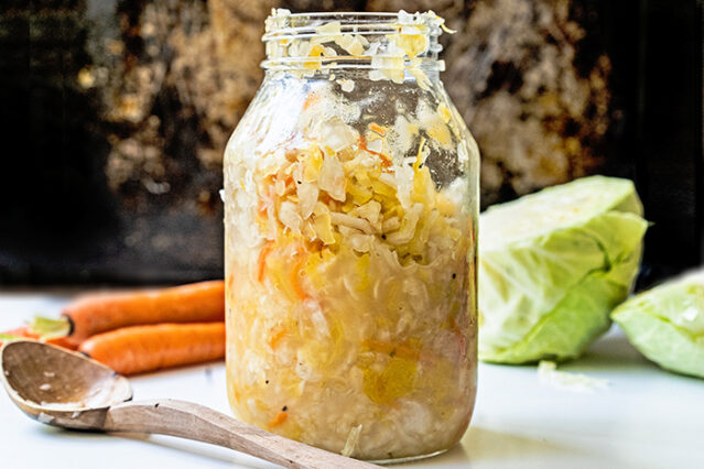 Large, open jar of sauerkraut in front of a head of cabbage and whole carrots.