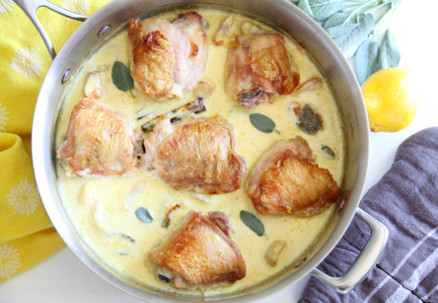 Chicken thighs in lemon sage cream sauce in pan with blue and yellow dish towels.