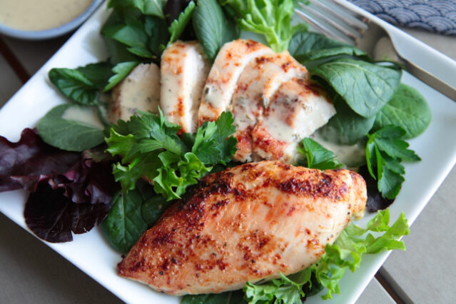 Whole and sliced chicken breast on a square white plate surrounded by greens.