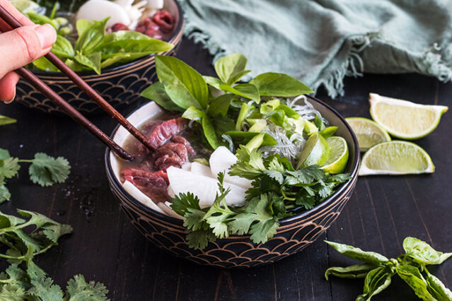 Hand using chopsticks to stir a bowl of beef pho with fresh herbs and lime wedges.