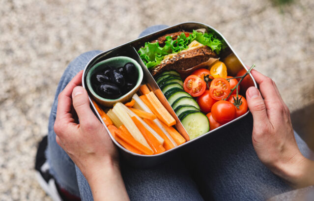 Over the shoulder view of a woman sitting on a bench holding a lunch box filled with assorted vegetables, olives, and half a sandwich.