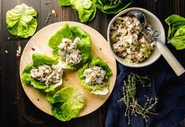 Four lettuce cups filled with chicken salad on a wooden serving tray. Bowl of chicken salad with a scoop. 