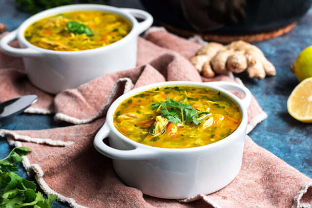 Two bowls of ginger turmeric chicken soup on pink cloth with fresh ginger in background.