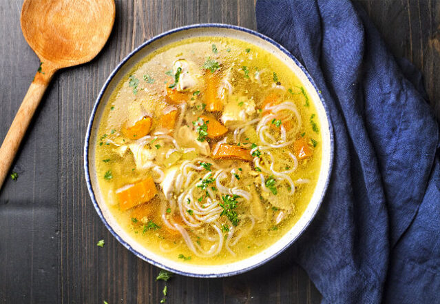 Overhead shot of chicken noodle soup with large wooden spoon and blue napkin.