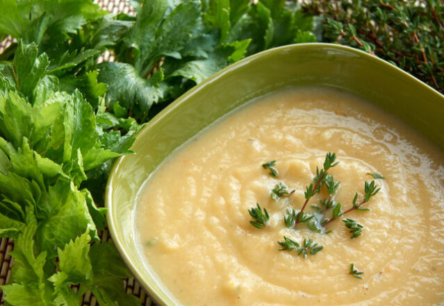 Square green bowl of blended celery root soup surrounded by fresh herbs.